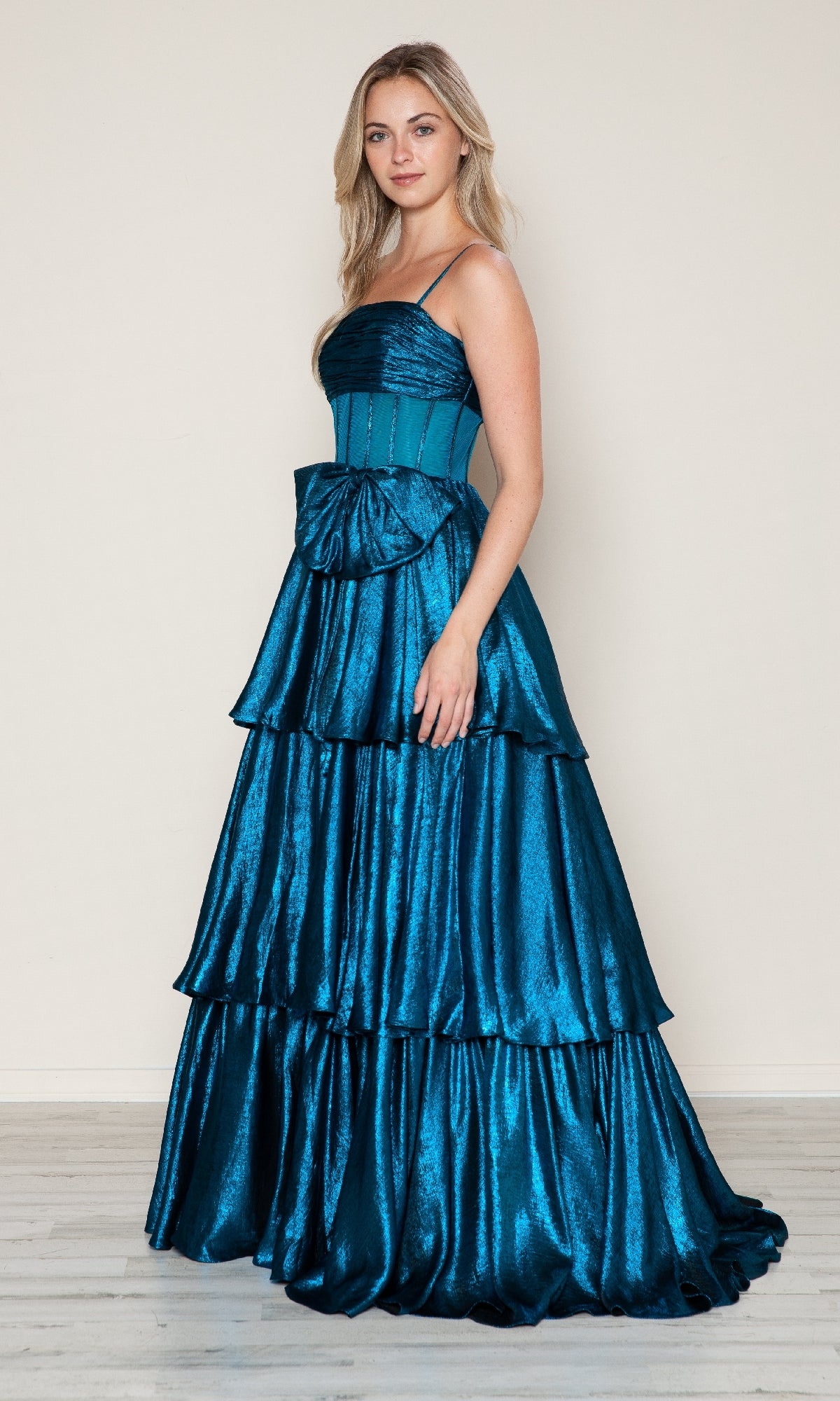 A woman poses against a plain beige wall in the Long Prom Dress: Poly USA 9718, a blue satin gown featuring an A-line silhouette, tiered skirt, thin straps, and a large waist bow.